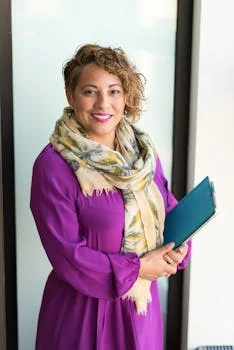 INÍCIO Smiling woman in a purple dress and scarf holding a tablet indoors.